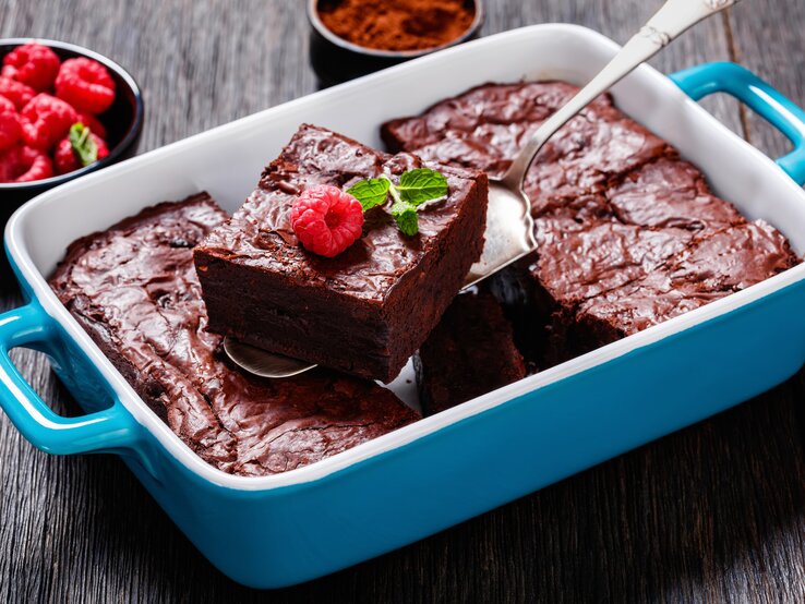 Himbeer-Brownies in blauer Auflaufform. Ein mit Himbeere und Minze belegtes Stück wird mit einem Tortenheber aus der Form gehoben. Daneben steht eine Schale mit Himbeeren. 