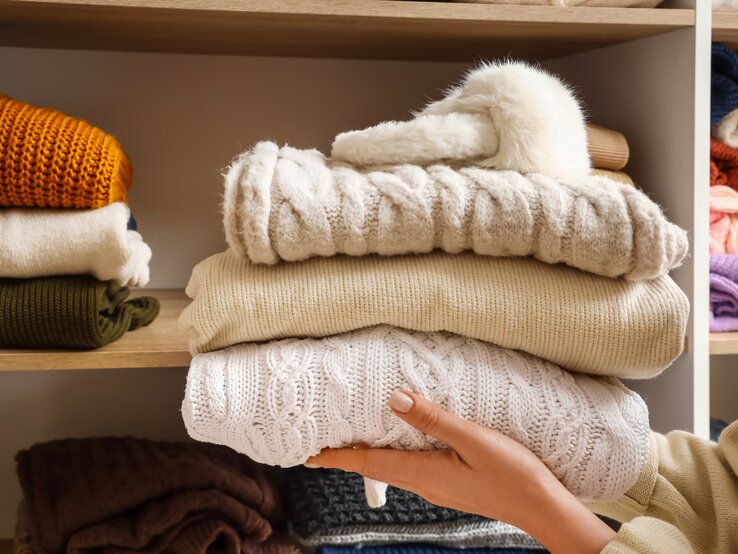Jemand hält ein paar Pullover vor einem Kleiderschrank.