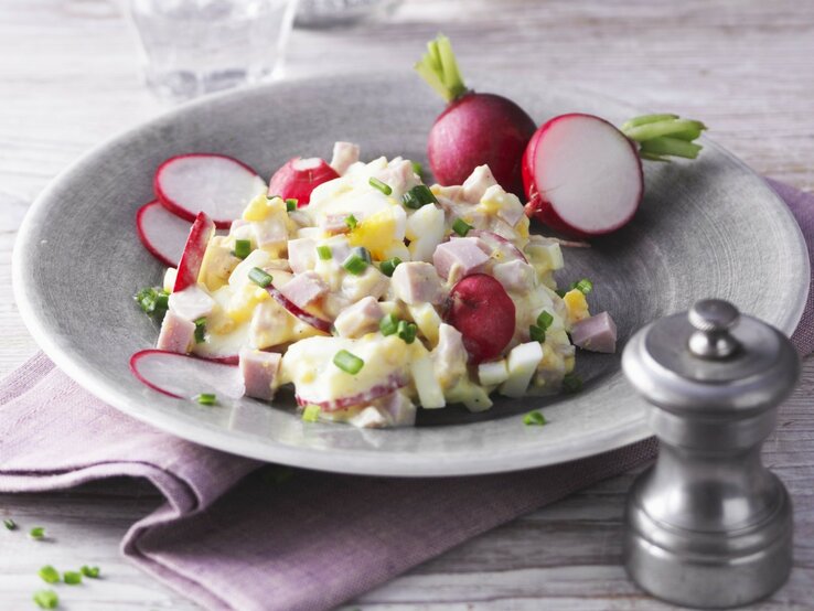 Eiersalat mit Pute und Radieschen in hellgrauer Schale auf lila Tuch und hellem Untergrund. Im Hintergrund ein Glas Wasser. Frontalaufnahme.