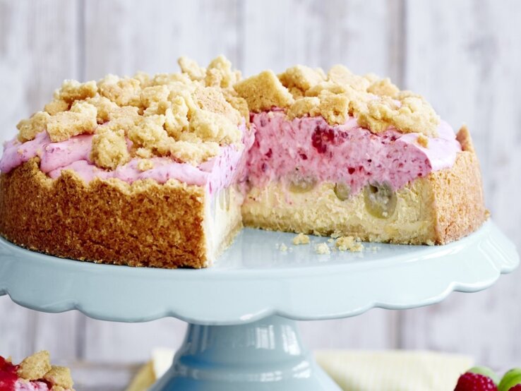 Auf einer Etagerie steht eine Stachelbeer-Quark-Streuseltorte. Sie ist mit einer rosa Creme gefüllt und angeschnitten. 