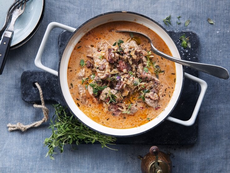 Eintopf mit Schweinefilet und getrockneten Tomaten, garniert mit frischem Thymian und angerichtet mit Sahne in einem großen Topf auf einer Schierferplatte. 