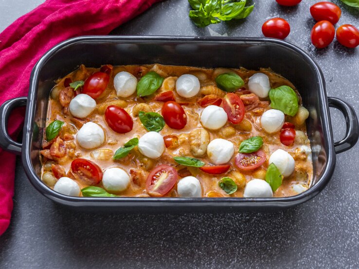 Schwarze Auflaufform gefüllt mit gebackenen Gnocchi mit Tomaten-Sahne-Soße und Mozzarella auf dunklem Untergrund. 