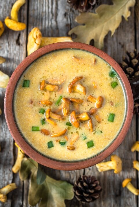 Aus der Vogelperspektive: eine Schüssel Kartoffelsuppe mit Pfifferlingen stehtauf einem Untergund aus Holz. Drumherum liegen Blätter, Tannenzapfen und frische  Pfifferling sowie eine Suppenkelle.