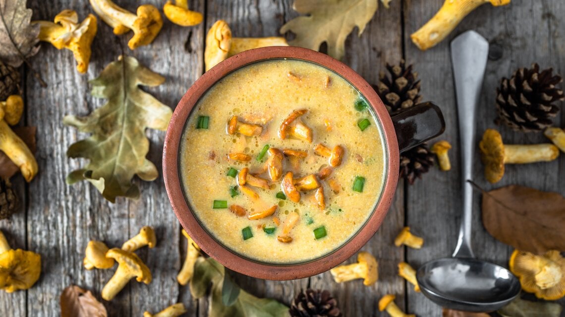 Aus der Vogelperspektive: eine Schüssel Kartoffelsuppe mit Pfifferlingen stehtauf einem Untergund aus Holz. Drumherum liegen Blätter, Tannenzapfen und frische  Pfifferling sowie eine Suppenkelle.