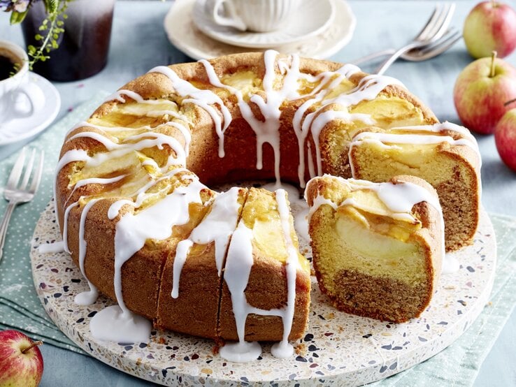 Angeschnittener Apfel-Kranzkuchen mit Zuckerguss auf Servierteller vor hellem Hintergrund mit Kaffeetassen und Äpfeln. 
