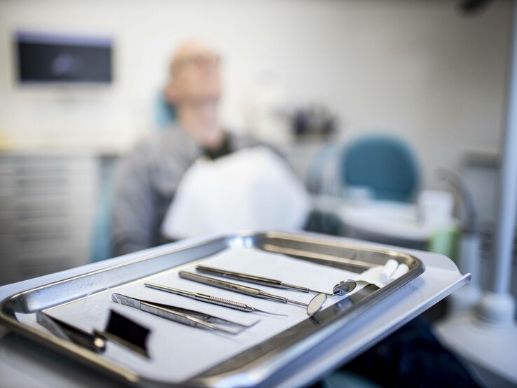 Im Hintergrund liegt eine Person auf einem Zahnarztstuhl. Im Vordergrund ist ein Tablett zu sehen.