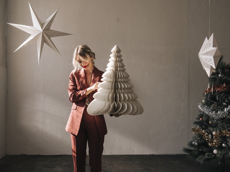 Schöne Frau, die das Wohnzimmer zu Weihnachten mit einem selbst gebastelten Tannenbaum aus Papier schmückt. 