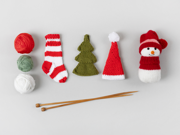 Auf einem hellen Untergrund sind gestrickte Weihnachtsdekorationen und Strickutensilien angeordnet. Von links nach rechts sieht man drei Wollknäuel in Rot, Grün und Weiß, daneben ein paar rot-weiß gestreifte, gestrickte Weihnachtssocken, einen grünen, gestrickten Tannenbaum, eine rote Weihnachtsmütze mit weißem Rand und Bommel, und einen gestrickten Schneemann mit roter Mütze und Schal. Im Vordergrund liegen zwei Holzstricknadeln.