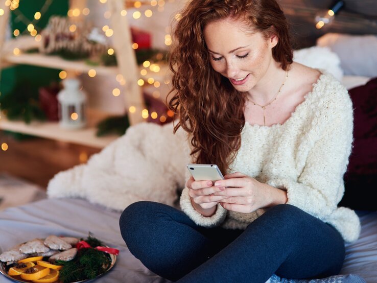 Junge Frau sitzt auf dem Sofa und verschickt Advent Sprüche an ihre Freunde und die Familie. Ihre Wohnung ist weihnachtlich dekoriert. 