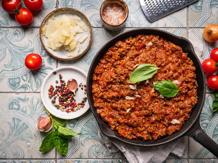 Gusseiserne Pfanne  mit Bolognese-Soße auf gekachelter Oberfläche. Umgeben ist der Topf von typischen Zutaten einer klassischen Bolognese. 