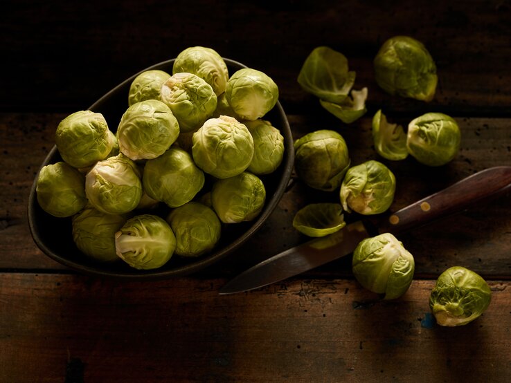 Rosenkohl  in dunkler Schale auf hölzernem Tisch, daneben ein Messer sowie einige angeschnittene Kohlköpfchen.