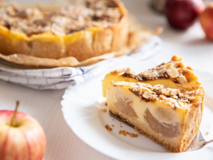 Angeschnittener Bratapfel-Schmandkuchen mit zwei einzelnen Stücken auf  einem weißen Teller. Der Kuchen präsentiert sich mit einer goldbraunen, mit Mandeln belegten Kruste.