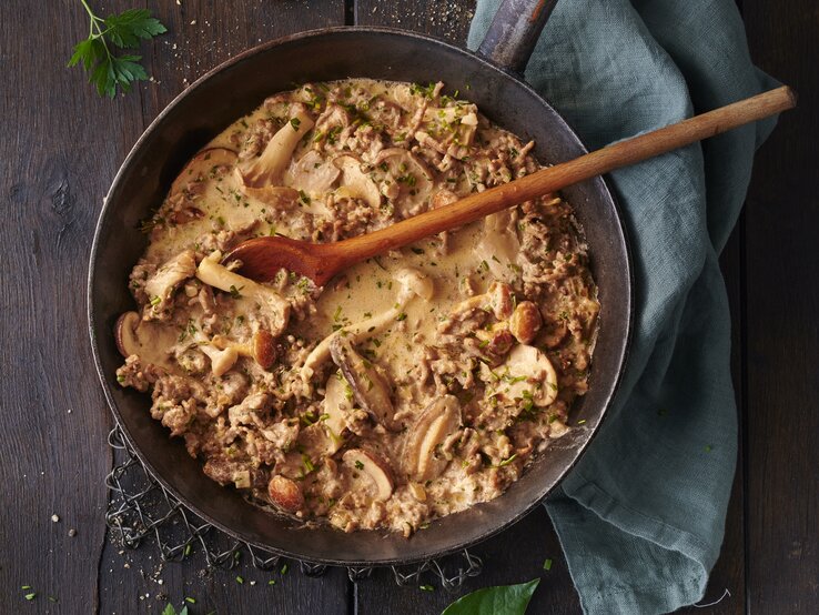 Hackfleisch-Jägerpfanne mit  gemischten Pilzen und frischen Kräutern. Das Gericht befindet sich in einer rustikalen Pfanne und besteht aus gebräuntem Hackfleisch gemischt mit einer Vielzahl von Pilzen, die in verschiedenen Größen und Formen zu sehen sind. Die Pilze sind goldbraun geröstet.