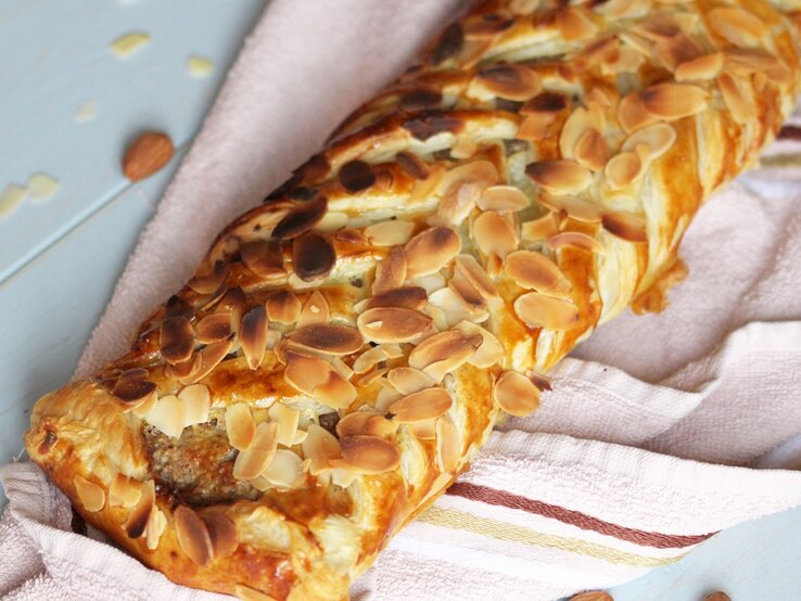 Ein Marzipan-Mandel-Strudel auf einem hellen Tisch mit rosa Tischdecke. Alles von schräg oben fotografiert.