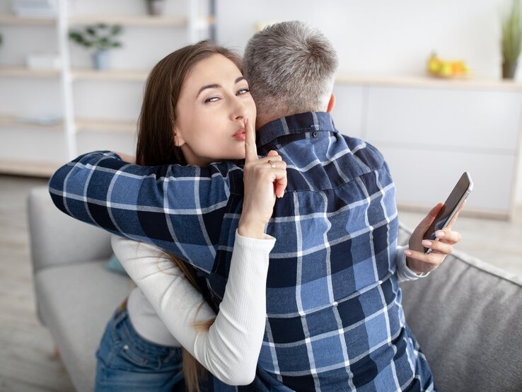 Pssst. Was Affären und Verhältnisse aber gemeinsam haben können, ist eine selbst gewählte Verheimlichung bzw. Heimlichkeit Frau und Mann umarmen sich und die Frau hat hinter dem Rücken des Mannes ein Handy in der Hand, guckt in die Kamera und macht das Hush-Zeichen.