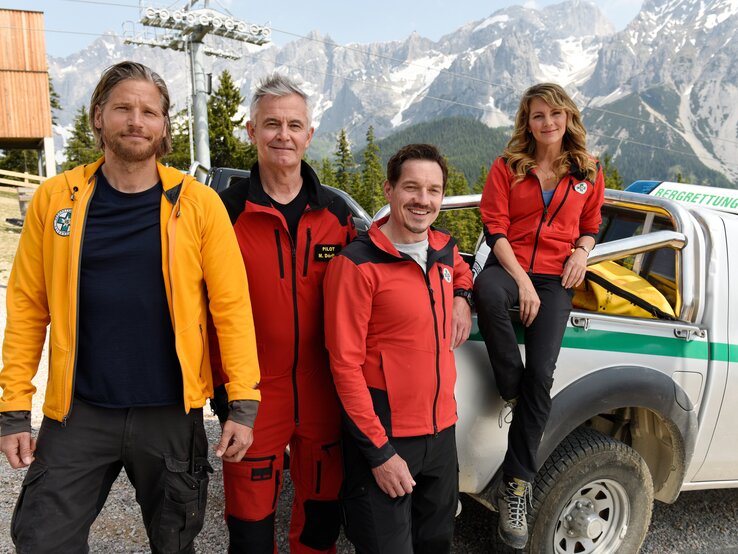 Vier "Die Bergretter"-Darsteller stehen in Rettungsdienstuniformen neben einem Offroad-Rettungsfahrzeug mit der Aufschrift "Bergrettung". Im Hintergrund sind Berggipfel zu sehen. Von links nach rechts: Sebastian Ströbel als Markus Kofler, Robert Lohr als Michael "Michi" Dörfler, Markus Brandl als Tobias Herbrechter und Luise Bähr als Katharina Strasser.