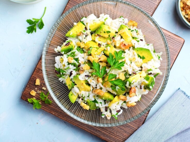 Draufsicht: Blauer Untergrund, ein quadratischtes Holzbrettchen, auf dem ein Teller aus Glas mit einem Avocado-Reissalat mit Mandeln liegt. Daneben liegt eine Schale mit ein paar gerösteten Mandeln.