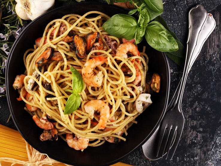 Ein schwarzer Teller auf grauem Tisch mit der Pasta. Daneben Kräuter, rohe Spaghetti und Besteck.