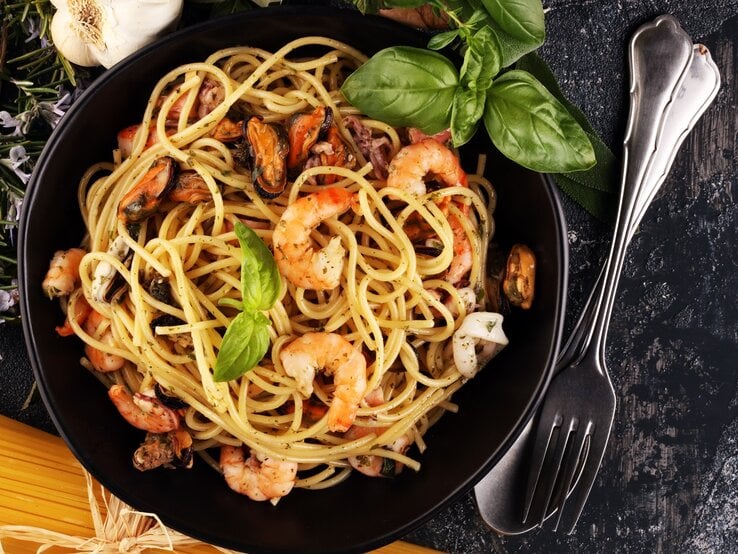 Ein schwarzer Teller auf grauem Tisch mit der Pasta. Daneben Kräuter, rohe Spaghetti und Besteck.