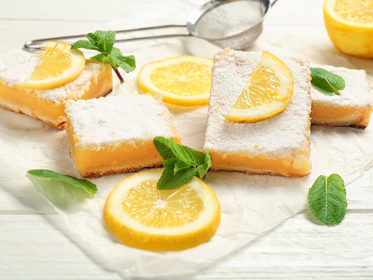 Frontalsicht: Auf einer weißen Platte aus Holz liegen mehrere Stücke Zitronenkuchen vom Blech, die mit Puderzucker bestäubt sind. Ebenfalls im Bild: Zitronenscheiben und Minzblätter.