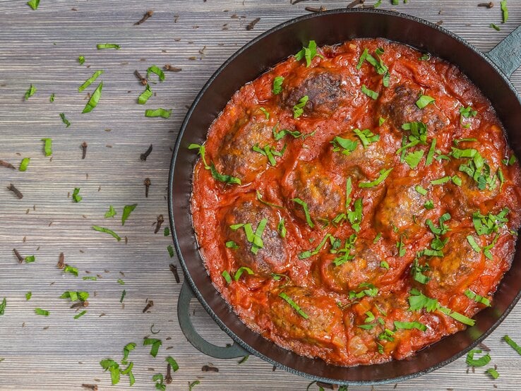 Soutzoukakia in Tomatensoße in schwarzer Pfanne auf Holztisch. 