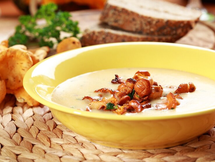 Ein gelber Suppenteller mit der Pfifferlingsuppe auf einer Bastmatte mit orange-gelbem Hintergrund. Außerdem Pfifferling, Brotscheiben und Petersilie im Hintergrund unscharf zu erkennen.