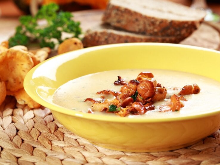 Ein gelber Suppenteller mit der Pfifferlingsuppe auf einer Bastmatte mit orange-gelbem Hintergrund. Außerdem Pfifferling, Brotscheiben und Petersilie im Hintergrund unscharf zu erkennen.
