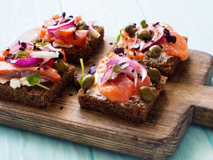 Vier Brote mit Lachs und Hüttenkäse auf einem dunklen Holzbrett und hellblauem Holzuntergrund von schräg oben fotografiert.