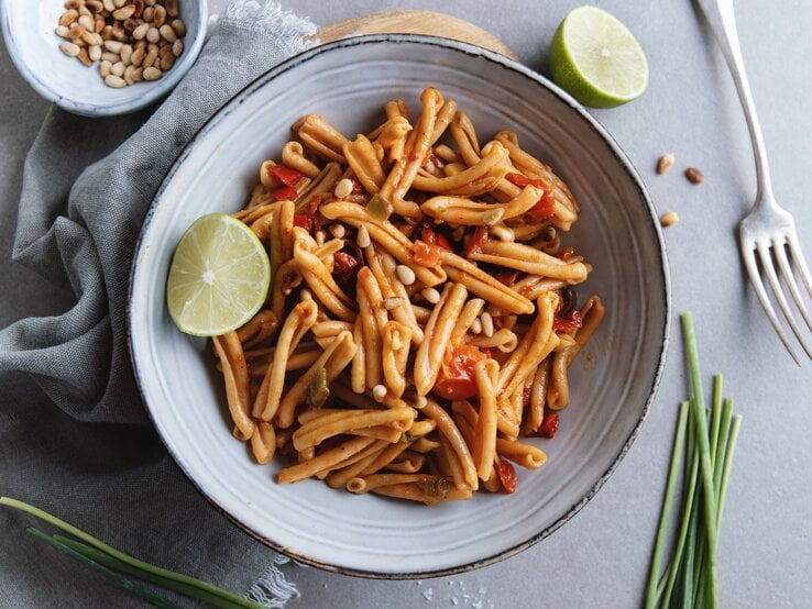 Tomaten-Pasta mit Pinienkernen dekoriert mit Limettenspalte auf weißem Teller. Daneben liegen Schnittlauch, Pinienkerne und eine Gabel. 