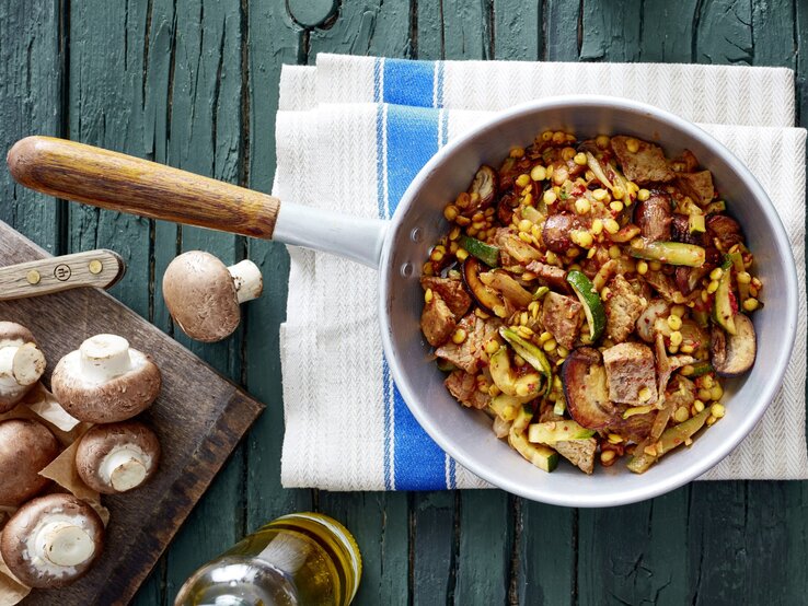 Schnitzel-Gemüse-Pfanne mit Linsen in Pfanne auf Küchentuch, daneben Flasche Öl und Champignons auf Brettchen, Untergrund dunkelgrünes Holz. 