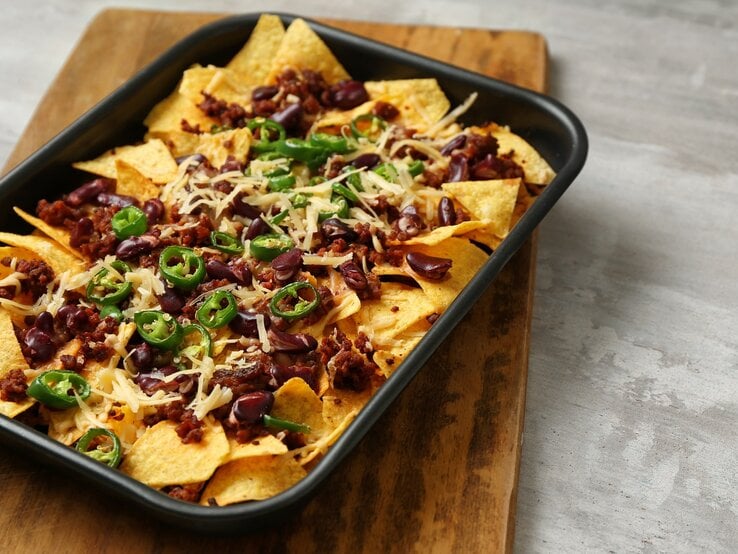 Eine schwarze Ofenform mit überbackenen Nachos mit Hack auf einem Holzbrett.