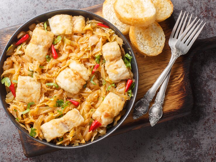 Sauerkrautauflauf mit Seelachs in schwarzer Schale auf Holzbrett. Daneben liegen Brotscheiben und Besteck.