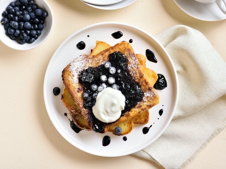 Macht richtig glücklich: French Toast mit Blaubeeren und Vanille-Schmand. Draufsicht: Ein weißer Teller mit French Toast mit Blaubeeren und Vanille-Schmand liegt auf einem hellen Untergrund. Daneben liegen ein helles, gobes Tuch sowie einen Schale mit frischen Heidelbeeren.