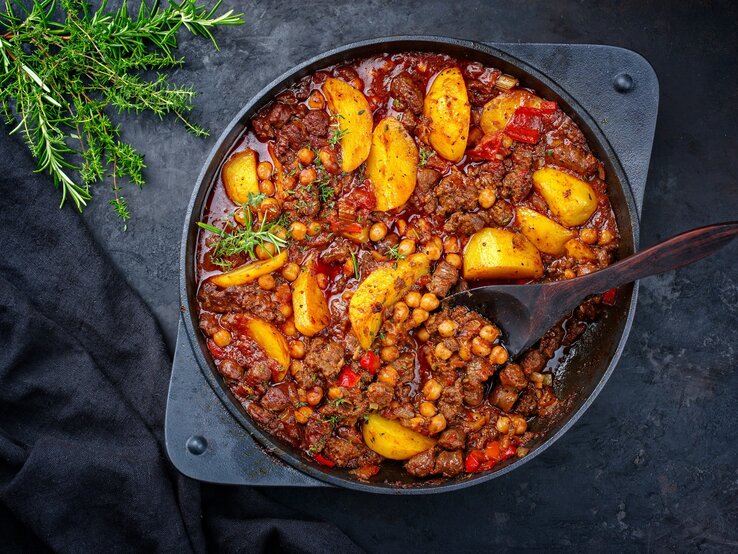 Draufsicht: Eine Hackfleisch-Pfanne mit Kichererbsen auf einem dunklen Untergrund. An der Seite liegen frische Kräuter.