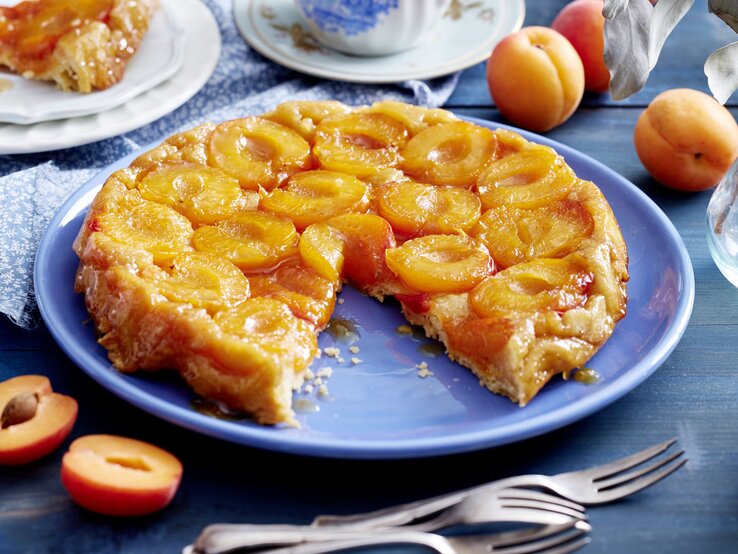 Aprikosen-Tarte-Tatin angeschnitten auf einer blauen Platte auf Holztisch mit Kaffeservice und ganzen Aprikosen