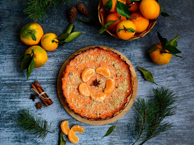 Ein Kürbiskuchen mit Mandarinen ist mit gehackten Pistazien und Mandarinen angerichtet. Der Kuchen steht auf einem blaugrauen Untergrund. Um den Kuchen herum liegen Zimt, Mandarinen ind Tannenäste als Dekoration.