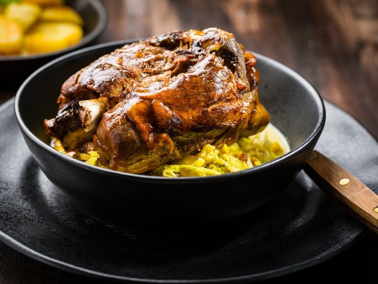 Frontal: Auf einem dunklen Untergrund stehen ein schwarzer Teller mit einer schwarzen Schüssel. Darin ist eine Bayerische Schweinshaxe auf sauerkarut.