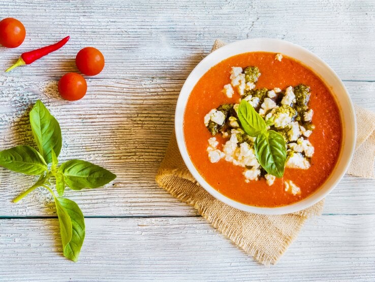 Rote Tomaten-Feta-Suppe mit Pesto und Basilikum in weüßer Schüssel auf Holz.