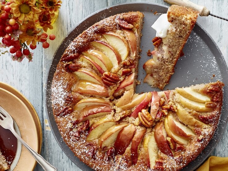 Ein grauer Teller mit dem Kuchen auf einem hellen Tisch direkt von oben fotografiert. Daneben rötliche Blumen und eine goldenes Tuch. 