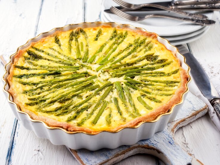 Eine vegane Spargelquiche in einer runden Quicheform auf weißem Holz, im Hintergrund Teller und Gabeln.