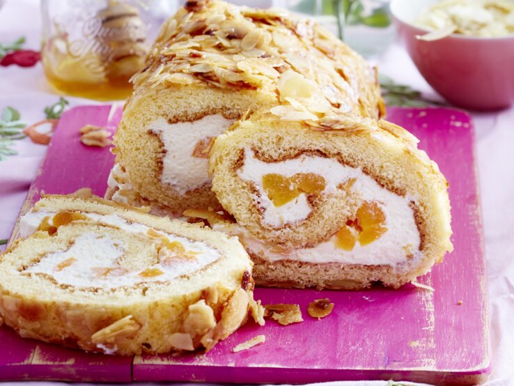 Lust auf einen Klassiker im neuen Gewand? Dann verpassen Sie nicht unser Rezept für eine Bienenstich-Biskuitrolle. Bienenstich-Biskuitrolle angeschnitten auf Holzbrett serviert.