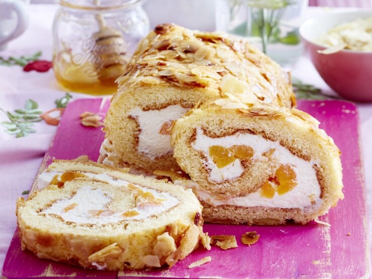 Lust auf einen Klassiker im neuen Gewand? Dann verpassen Sie nicht unser Rezept für eine Bienenstich-Biskuitrolle. Bienenstich-Biskuitrolle angeschnitten auf Holzbrett serviert.