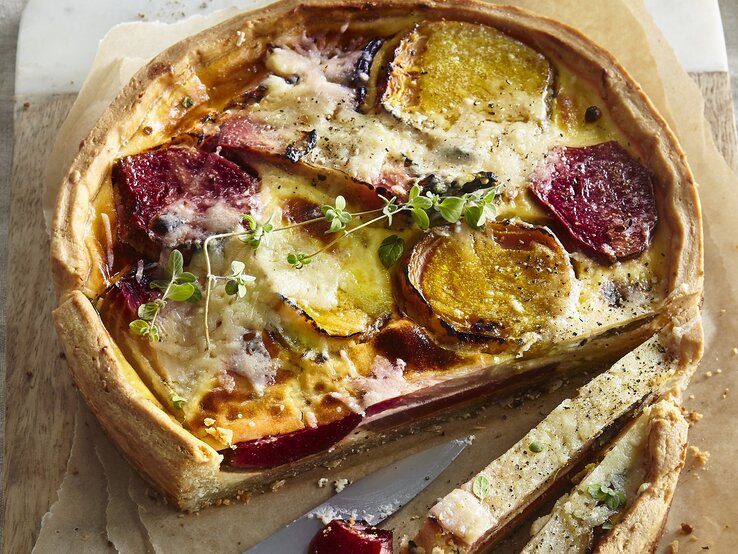 Angeschnittene Quiche mit Thymianzweig, geschmolzenem Käse auf Backpapier und geteiltem Holzbrett mit Steingutrand aus der Vogelperspektive. Rote Bete und Gelbe Bete in Scheiben sichtbar. Der Rand der Quiche leicht erhöht und gebräunt. Zwei Scheiben längs aufgeschnitten. Messer mit silberner Klinge und Holzgriff zwischen den Scheiben aufgelegt.