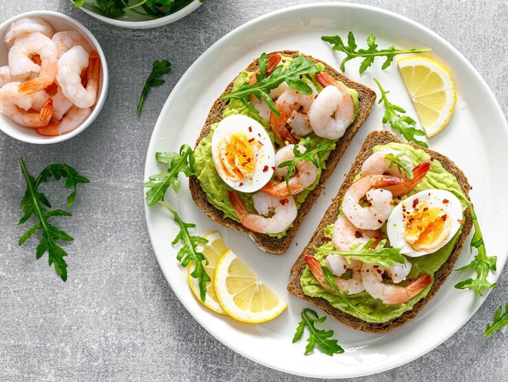 Ein weißer Teller mit den belegten Brotscheiben auf einem grauen Steintisch mit einem Schälchen mit Garnelen und einem mit Rucola direkt von oben fotografiert.