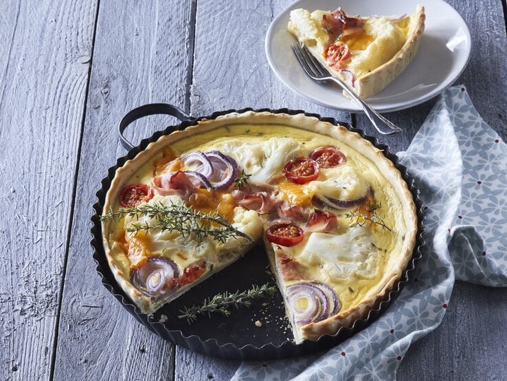 Eine Backform mit der frisch gebackenen Blumenkohl-Quiche mit Schinken und Räucherlachs neben einem Teller auf dem ein Stück der Quiche mit einer Gabel liegt, vor einem hellblauem Hintergrund.