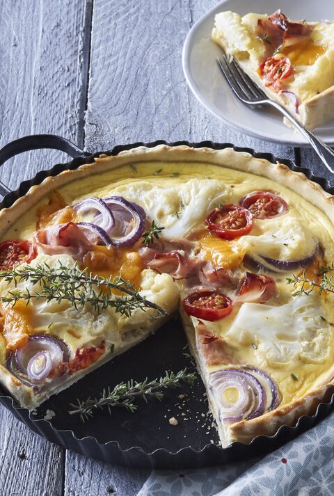 Eine Backform mit der frisch gebackenen Blumenkohl-Quiche mit Schinken und Räucherlachs neben einem Teller auf dem ein Stück der Quiche mit einer Gabel liegt, vor einem hellblauem Hintergrund.