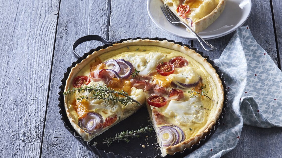 Eine Backform mit der frisch gebackenen Blumenkohl-Quiche mit Schinken und Räucherlachs neben einem Teller auf dem ein Stück der Quiche mit einer Gabel liegt, vor einem hellblauem Hintergrund.