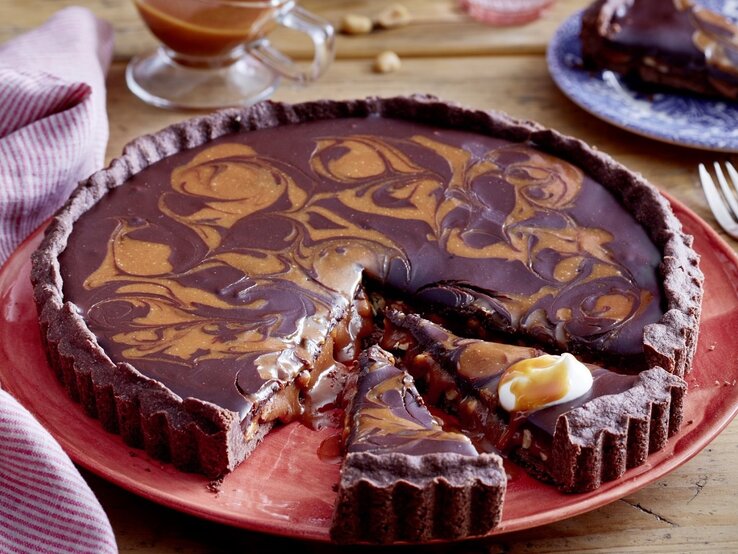 Diese Erdnuss-Karamell-Marmortarte zergeht förmlich auf der Zunge. Das ist mal wieder ein richtig leckerer Schokokuchen! Auf einem Teller liegt eine Erdnuss-Karamell-Marmortarte. Sie ist mit Karamell in einem Muster überzogen und ist extra cremig.