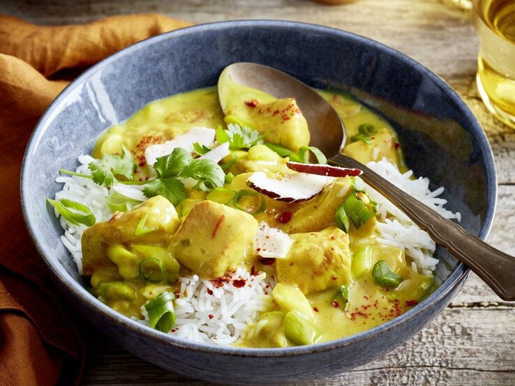 Eine Schüssel mit Basmati-Reis und cremigem Curry mit Hähnchenstücken, garniert mit frischen Frühlingszwiebeln, Koriander, Chiliflocken und Kokosraspeln. Eine Gabel liegt am Schüsselrand, im Hintergrund ist ein Getränk zu sehen.