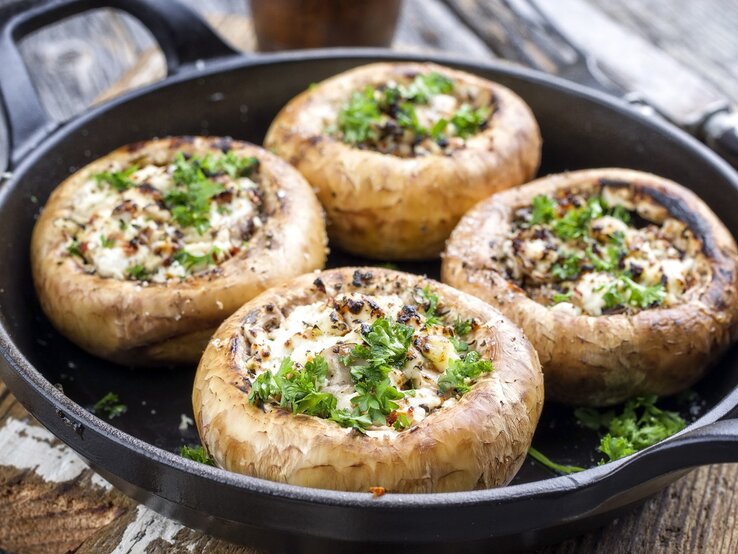 Eine gusseiserne Pfanne mit vier gefüllten Champignons auf einem dunklen Holztisch von schräg oben fotografiert.